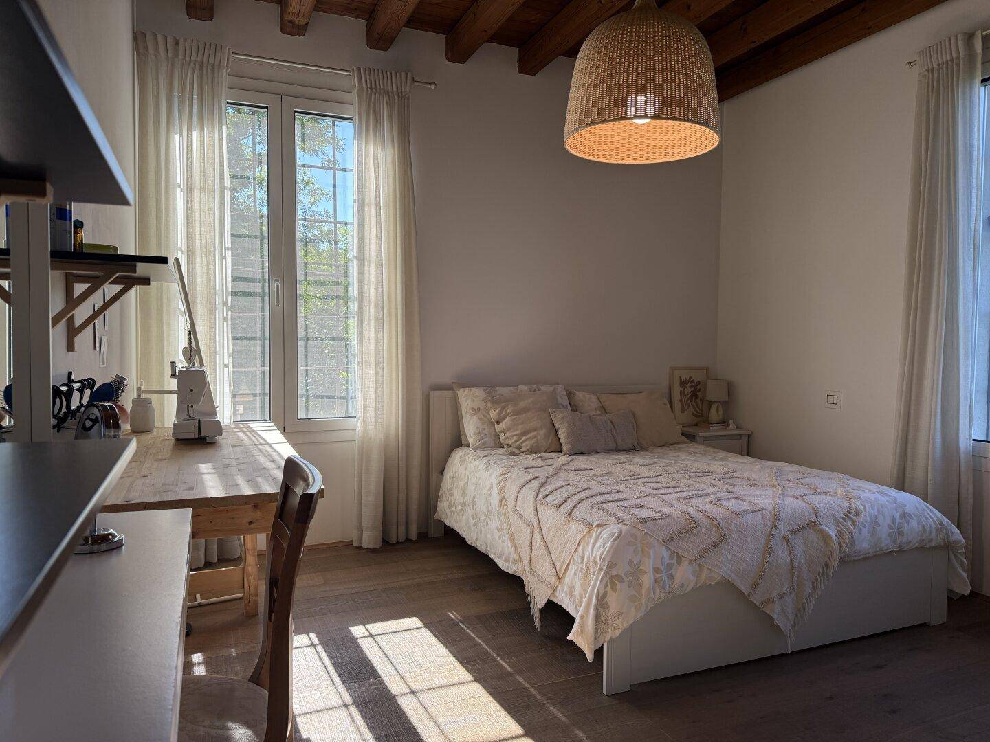 Helles Schlafzimmer mit Holzbalkendecke, Parkettboden und einem Arbeitsbereich am Fenster.