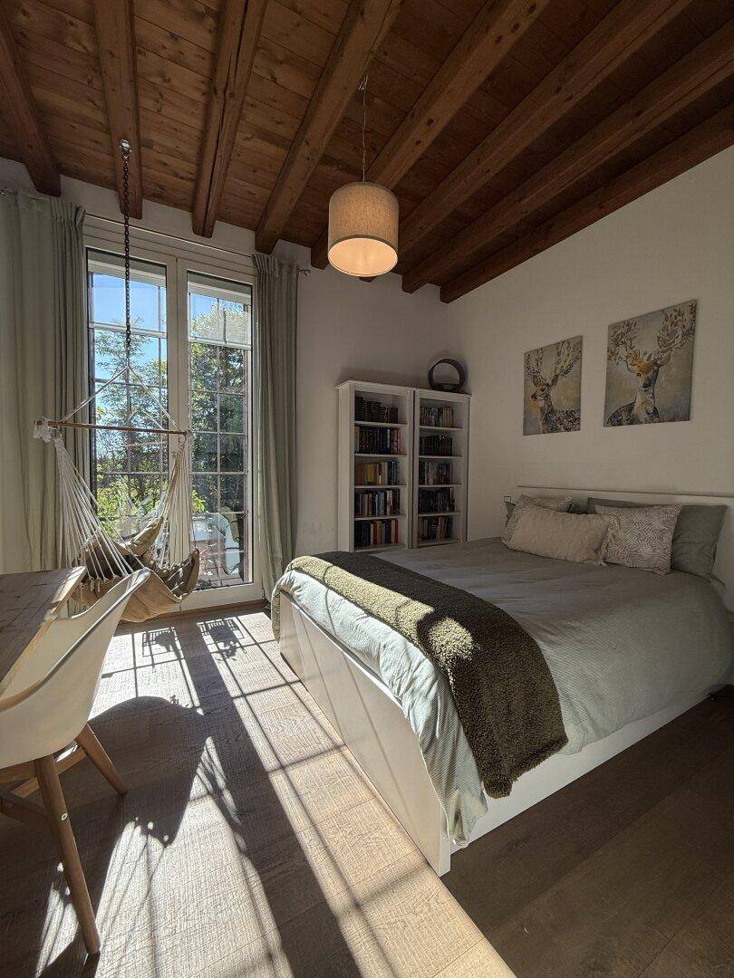 Gemütliches Schlafzimmer mit Holzbalkendecke, viel Tageslicht und einem Hängesessel am Fenster.