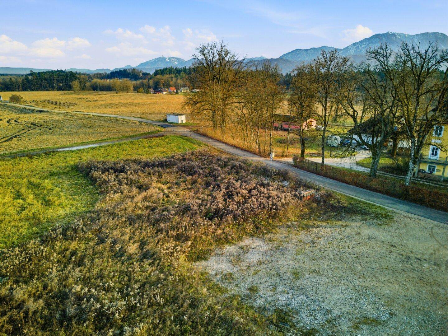 Weitläufige Landschaftsaufnahme mit Feldern, Bäumen und einer Straße, im Hintergrund sind Berge zu sehen.