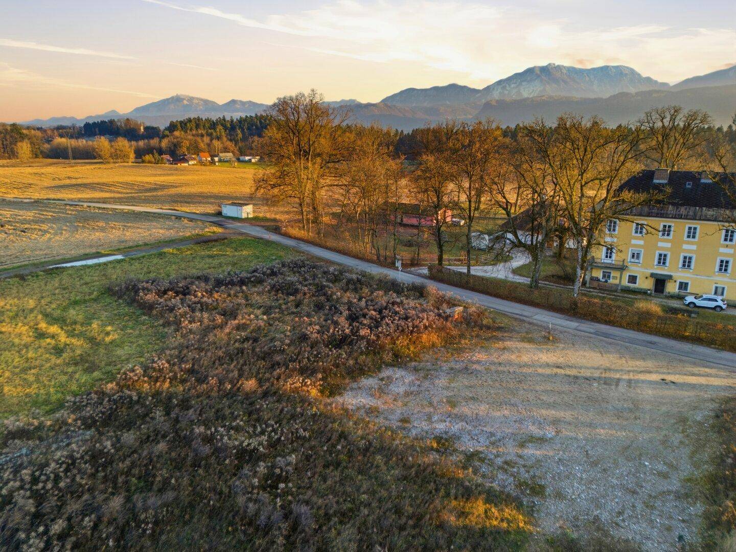 Ländliche Szenerie bei Sonnenuntergang mit Feldern, Bäumen und einem gelben Gebäude, Berge im Hintergrund.