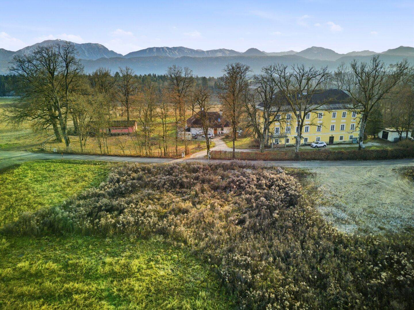 Weitläufige Landschaft mit einem markanten gelben Gebäude, umgeben von Feldern und Bäumen, mit Bergen am Horizont.