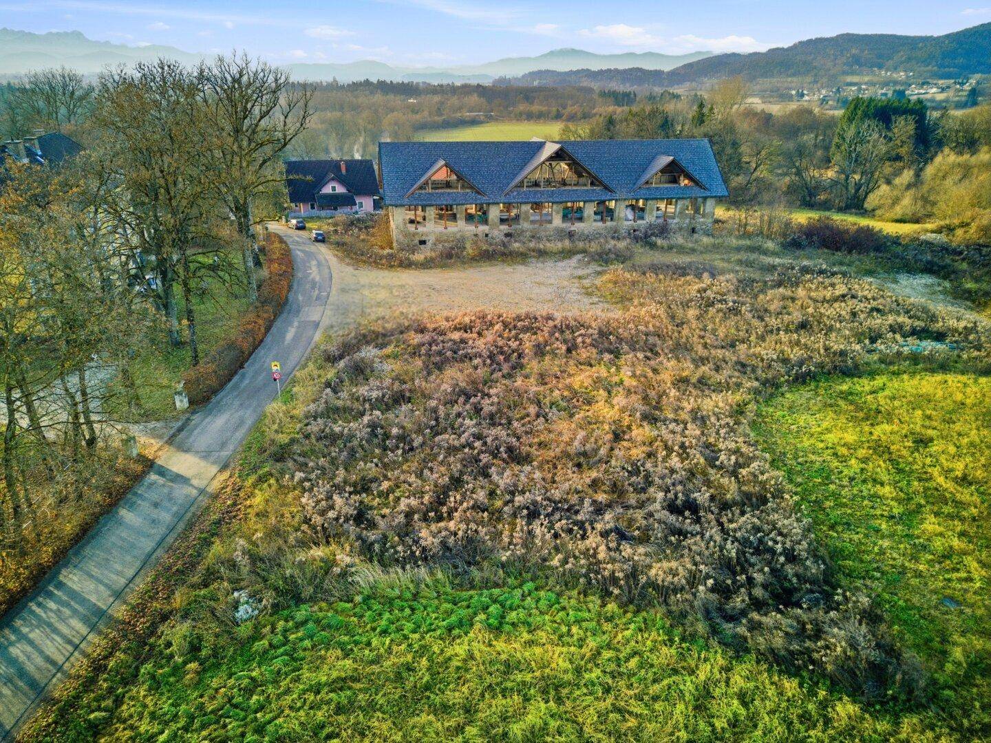 Luftaufnahme eines großen Gebäudes mit markantem Dach, umgeben von Feldern und Bäumen, mit Bergen im Hintergrund.