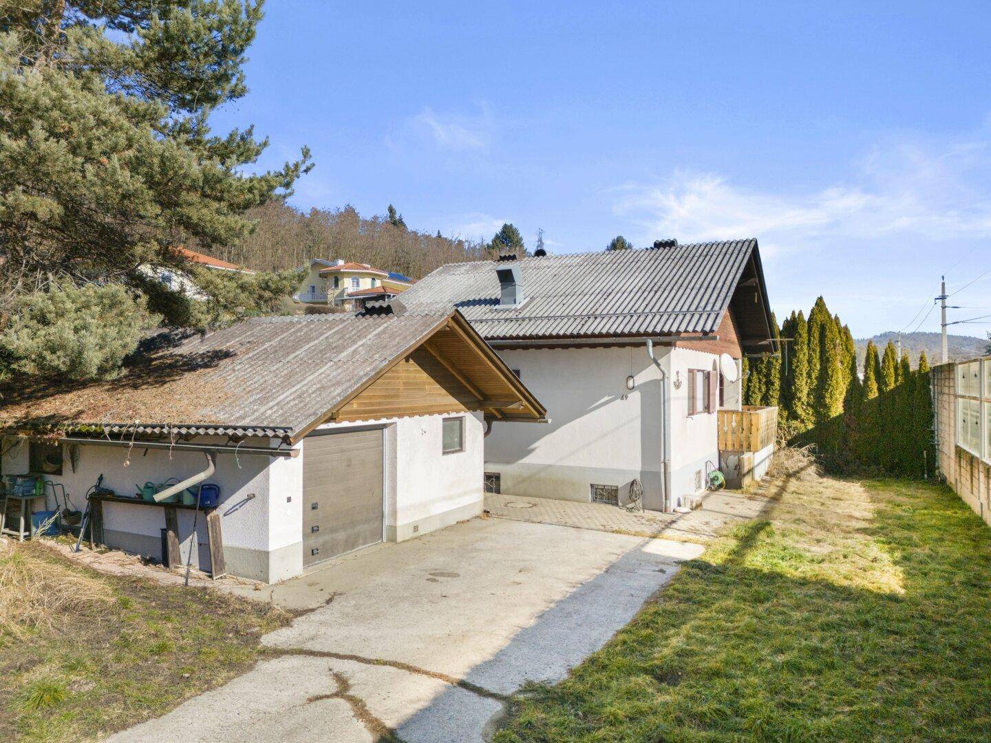 Hausfassade mit Garage und Gartenbereich, umgeben von hohen Hecken und Bäumen im Sonnenlicht.
