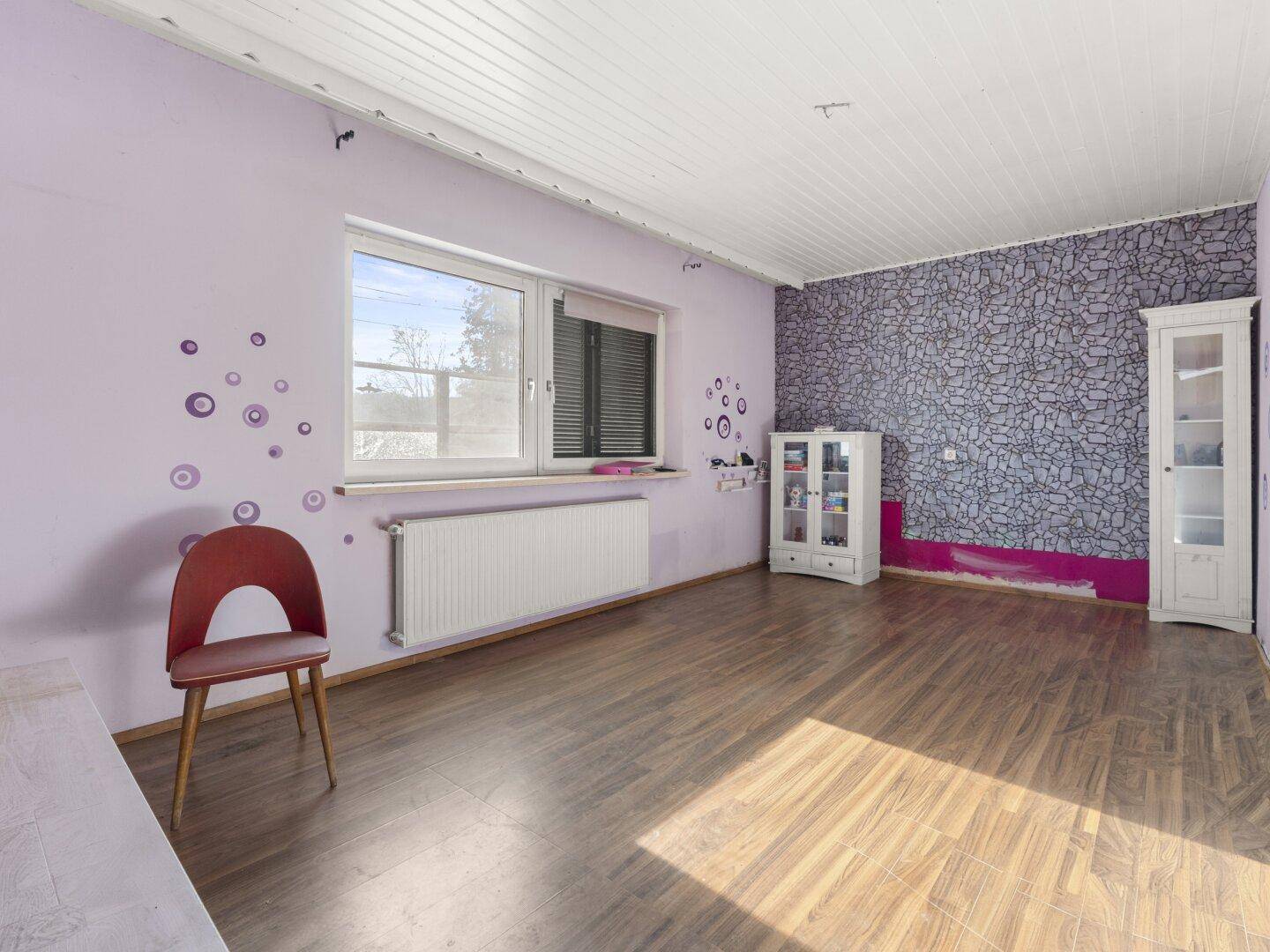 Helles Zimmer mit Laminatboden, lila Wänden und gemusterter Tapete, gut beleuchtet.
