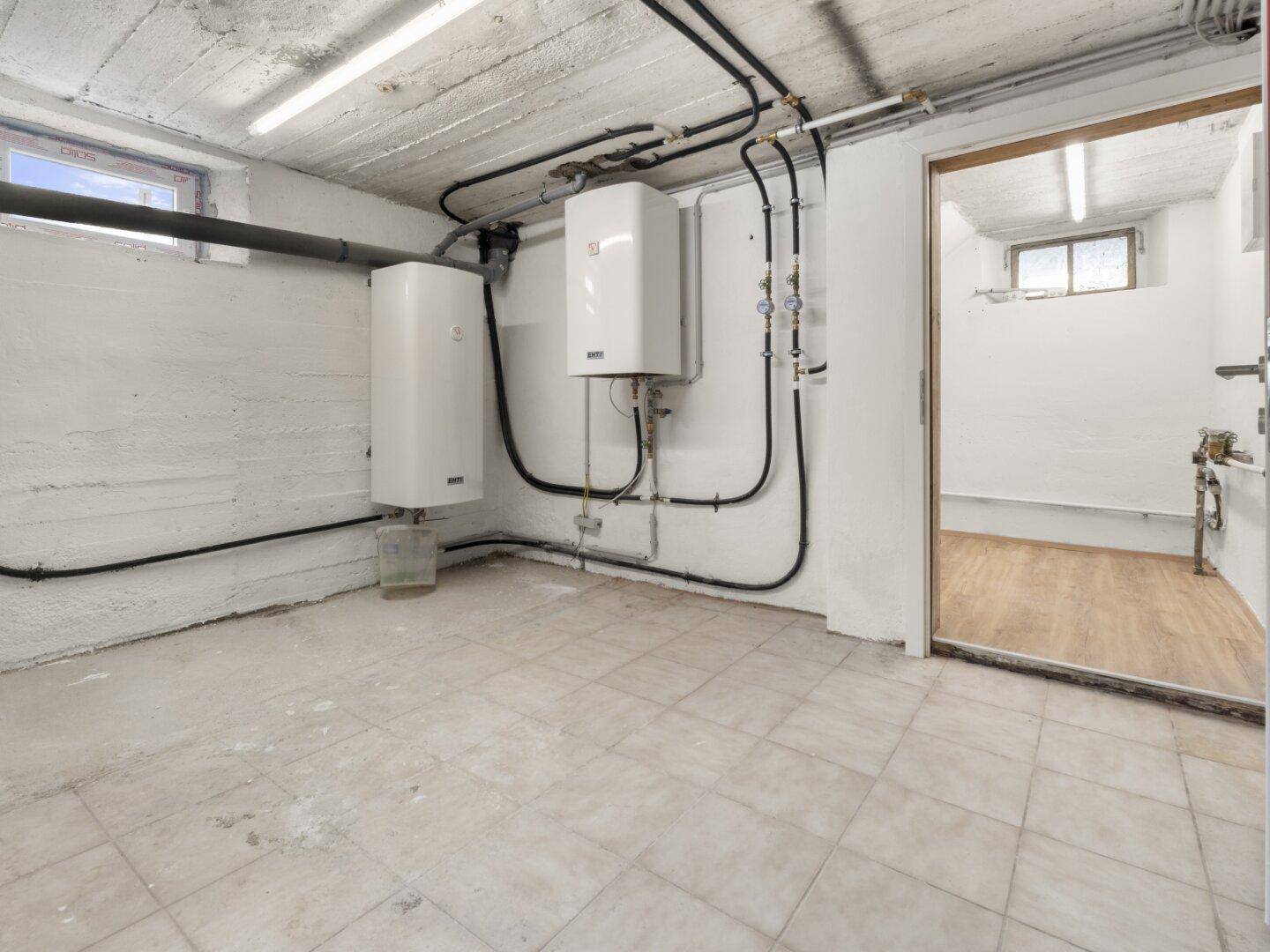 Technikraum im Keller mit Warmwasserboilern, sichtbaren Leitungen und Fliesenboden.