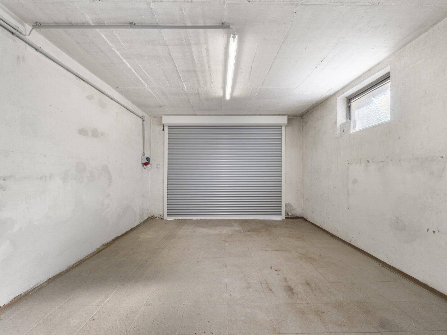 Geräumige Garage mit Rolltor, Betonboden und einem kleinen Fenster für Belüftung.