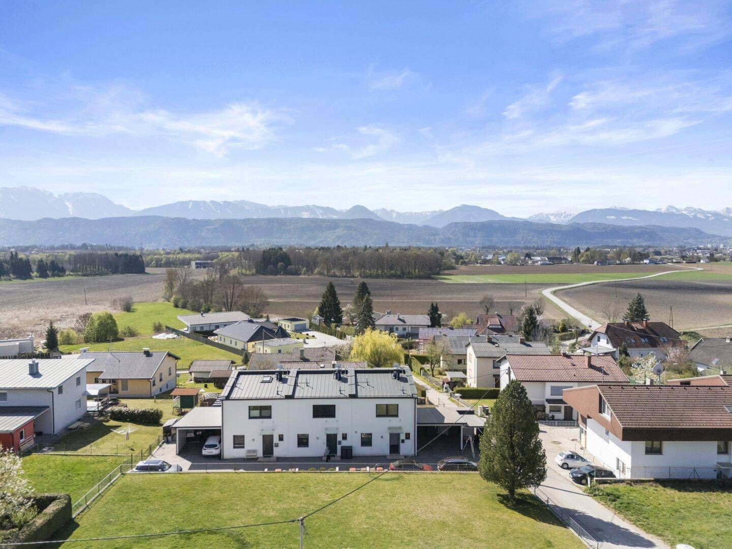 Weitläufige Luftaufnahme einer Wohnsiedlung mit grünen Feldern und majestätischem Bergpanorama im Hintergrund.