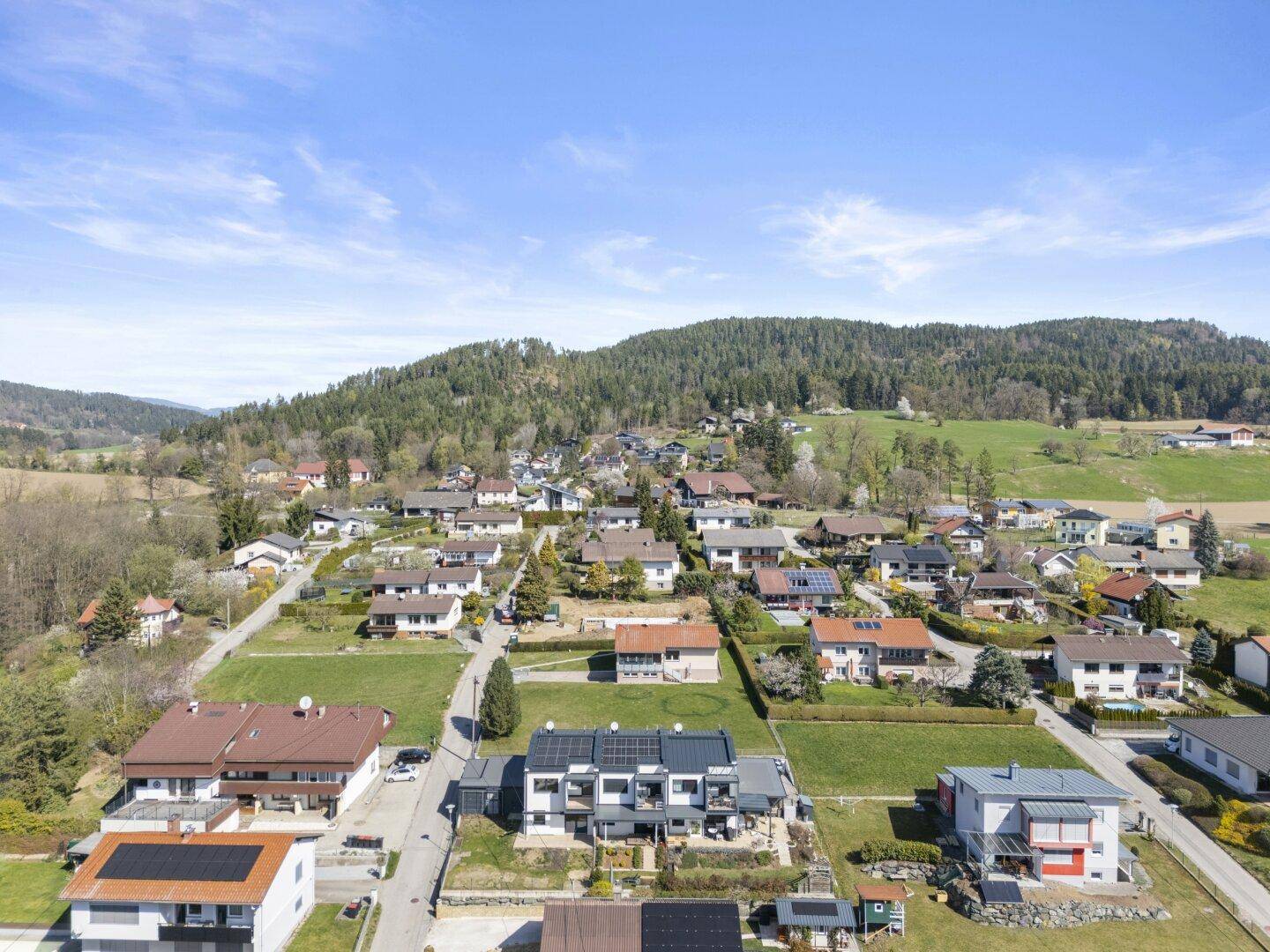Luftaufnahme einer Wohngegend mit modernen Häusern, umgeben von grünen Hügeln und Wäldern unter blauem Himmel.