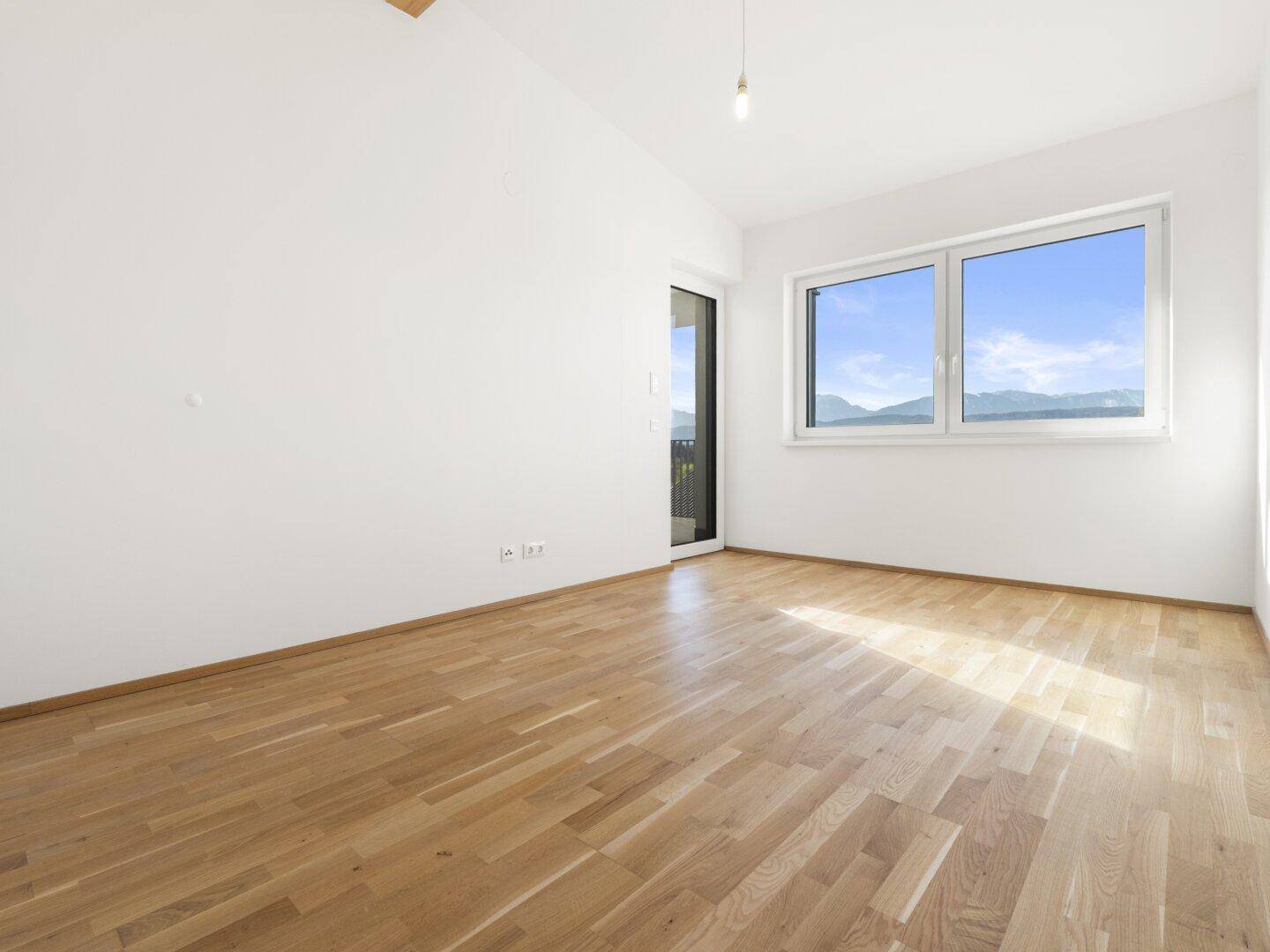 Heller, unmöblierter Raum mit Holzparkettboden, großen Fenstern und Zugang zum Balkon.