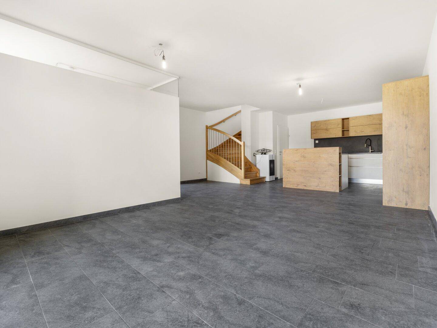 Weitläufiger, unmöblierter Wohnbereich mit Fliesenboden und Zugang zur Küche und Treppe.