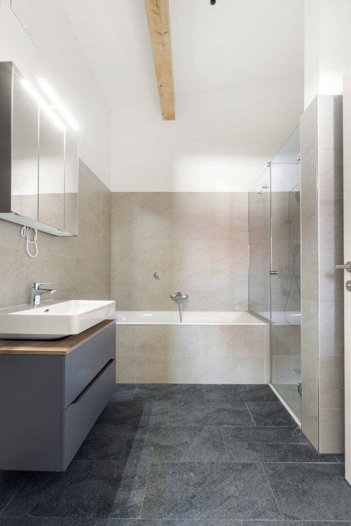 Helles Badezimmer mit modernen Armaturen, Badewanne, Dusche und sichtbarem Deckenbalken.