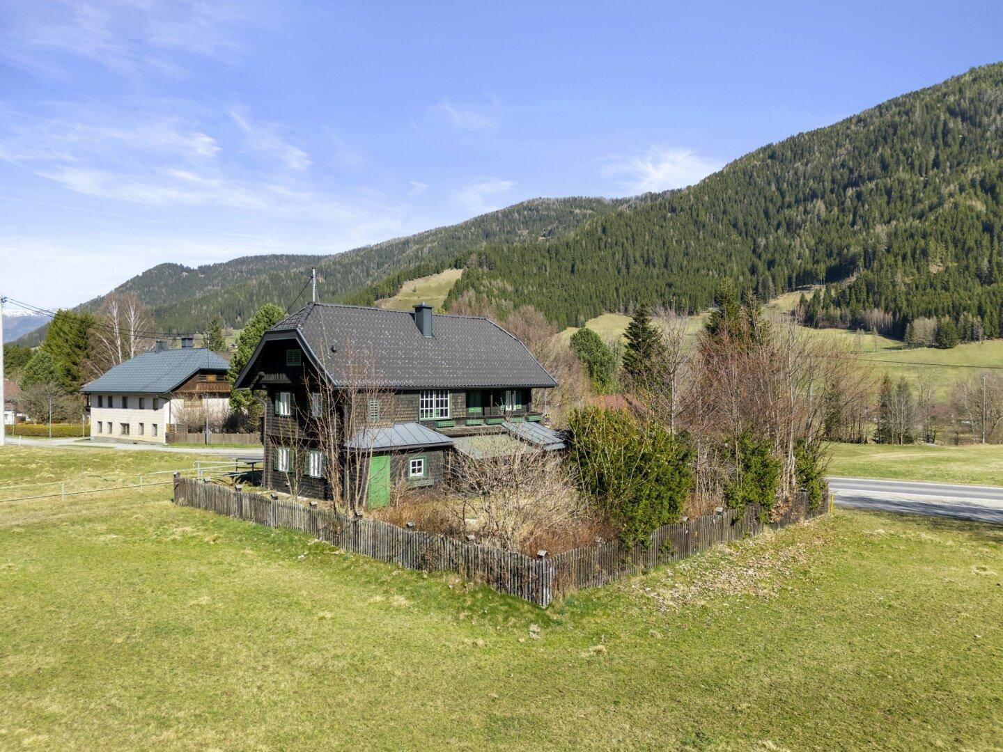 Traditionelles Holzhaus in ländlicher Umgebung mit grünen Wiesen und bewaldeten Bergen im Hintergrund.