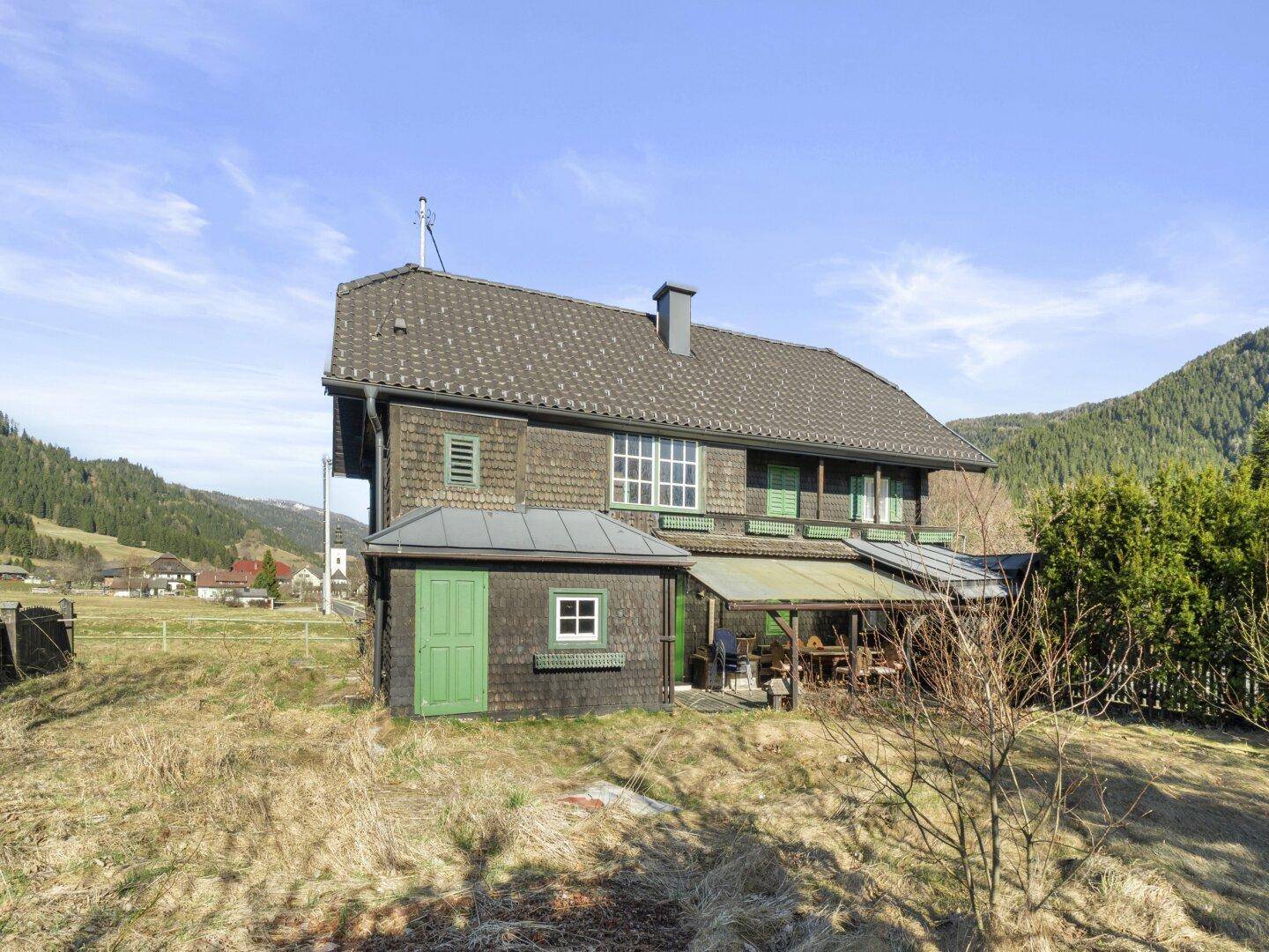 Rückansicht des Holzhauses mit Anbau und überdachtem Bereich, umgeben von Wiesen.