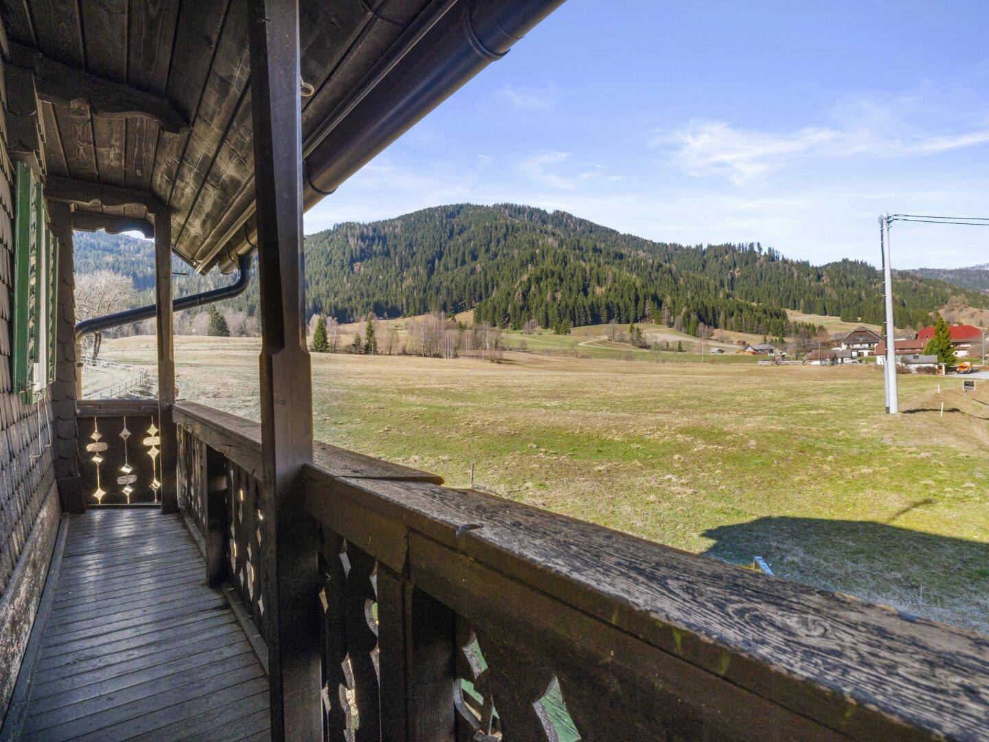 Holzbalkon mit traditionellem Geländer und freiem Blick auf weite Wiesen und Berge.