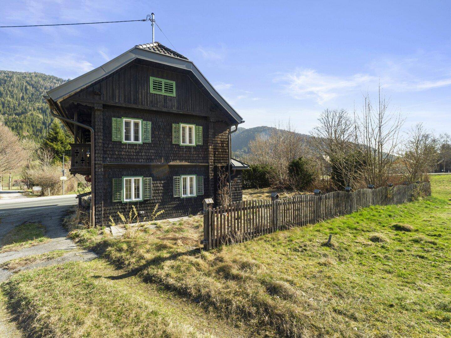 Seitenansicht des traditionellen Holzhauses mit Holzzaun und weiten Wiesenflächen.