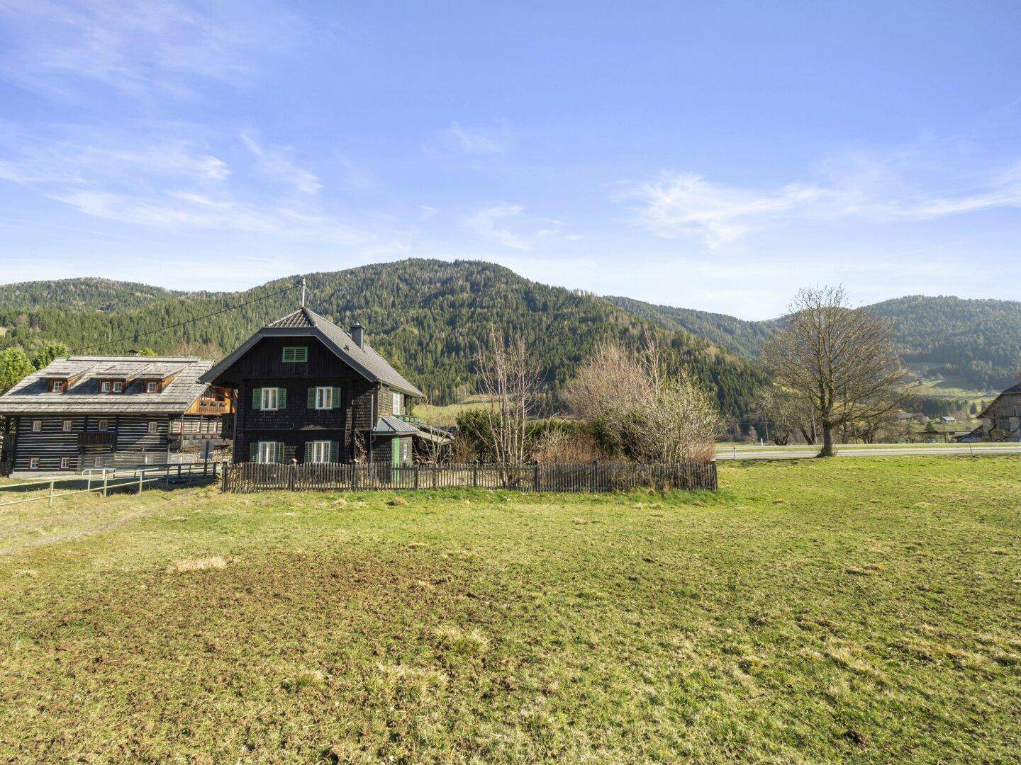 Das traditionelle Holzhaus eingebettet in eine weite, grüne Wiesenlandschaft mit Bergen.