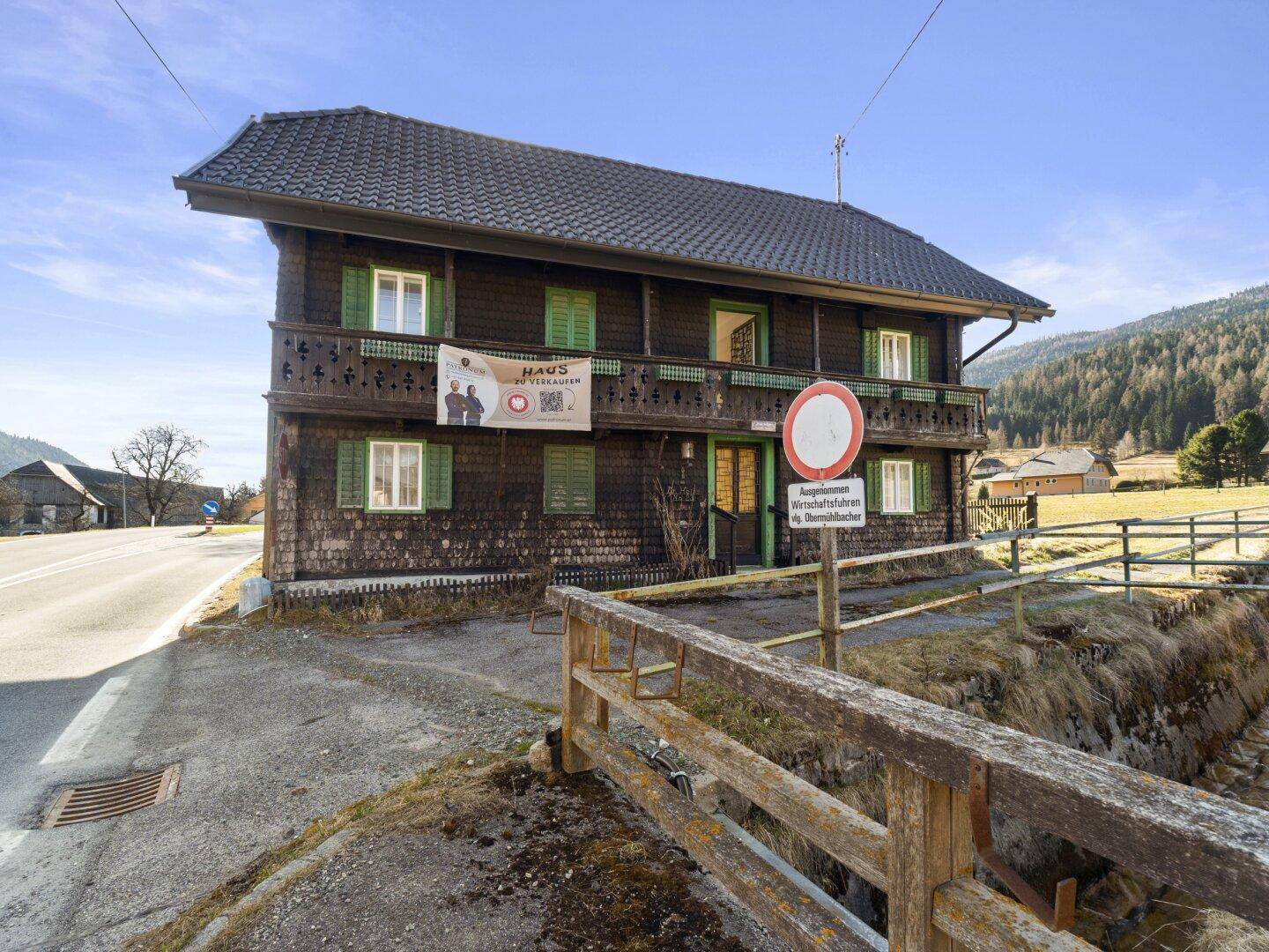 Traditionelles Holzhaus mit Balkon und Werbebanner an der Fassade, direkt an einer Straße gelegen.