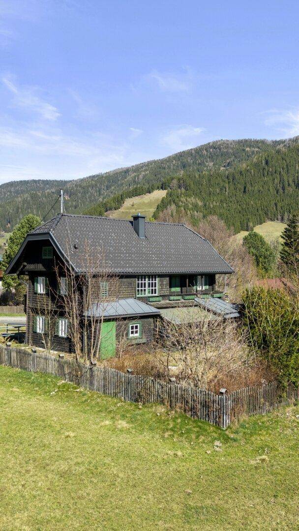 Traditionelles Holzhaus mit dunkler Fassade und grünen Fensterläden vor einer Bergkulisse.