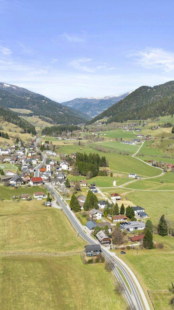 Weitläufige Luftaufnahme eines Dorfes in einem grünen Tal, umgeben von bewaldeten Bergen.