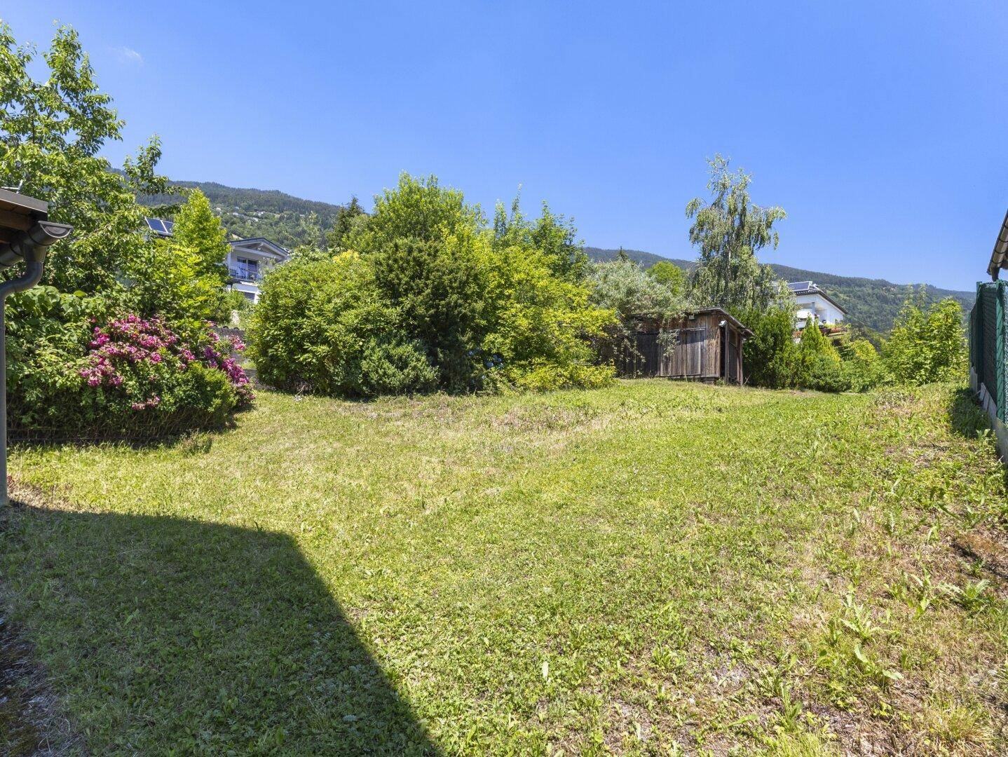 Sonniger Gartenbereich mit Rasenfläche, Büschen und einem kleinen Holzschuppen im Hintergrund.