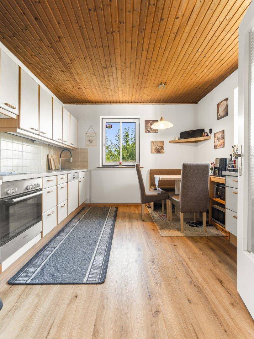 Helle Wohnküche mit weißer Einbauküche, Holzbalkendecke und Essbereich am Fenster.