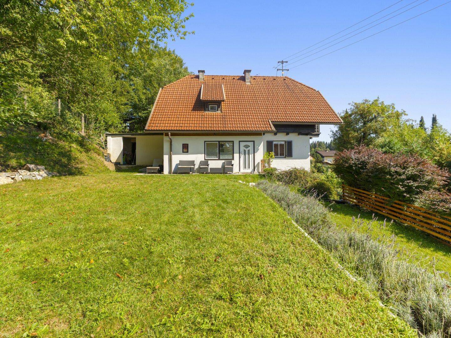 Vorderansicht des Hauses mit kleiner Terrasse und Rasenfläche, umgeben von Bäumen.