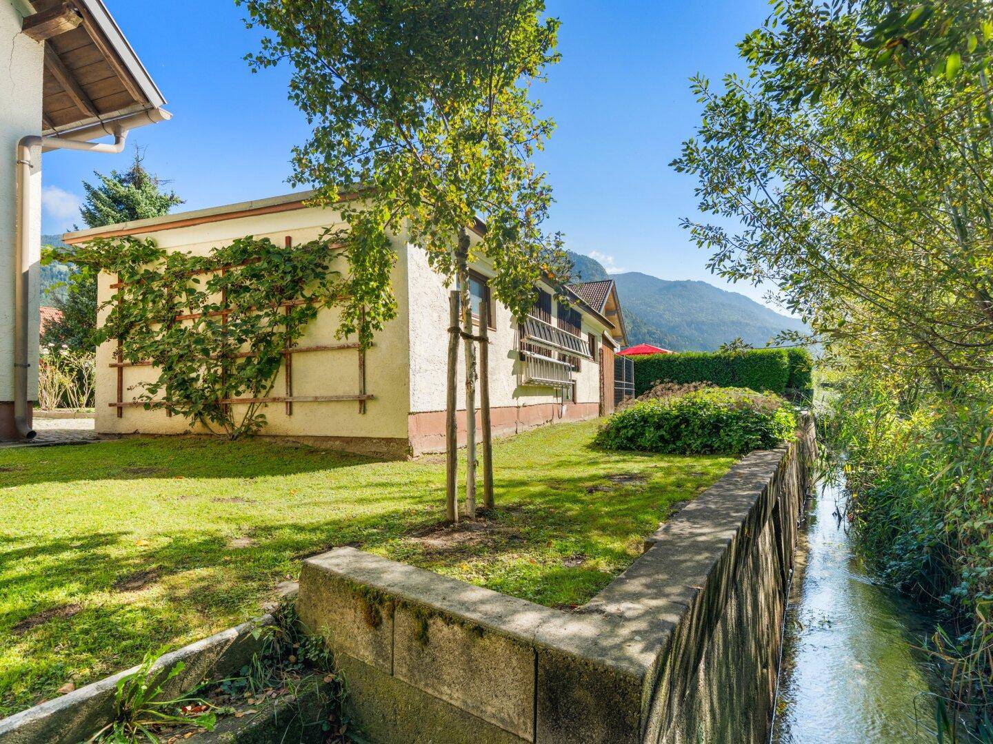 Seitenansicht des Hauses mit begrüntem Bereich und einem kleinen Bachlauf am Grundstücksrand.
