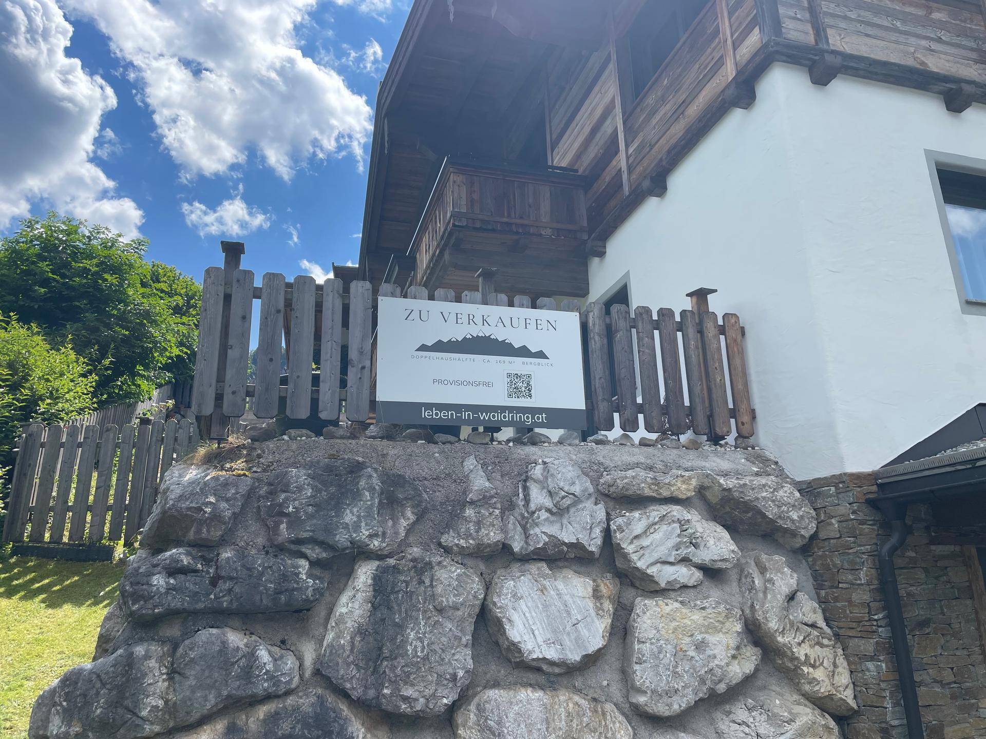 Chalet-ähnliches Gebäude mit traditioneller Holzfassade und einem 'Zu Verkaufen'-Schild auf einer Steinmauer.