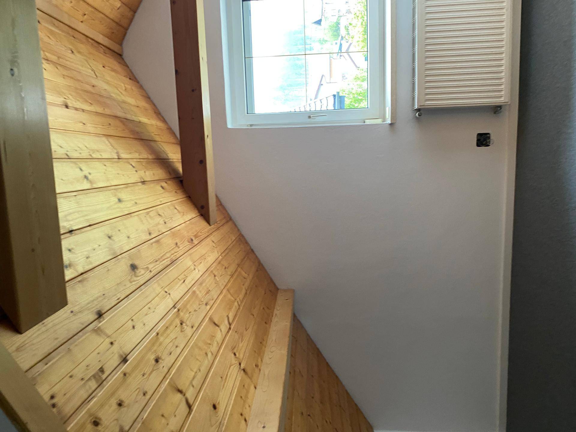 Holztreppe führt in ein helles Obergeschoss mit Fenster und Heizkörper, umgeben von Holzverkleidung.