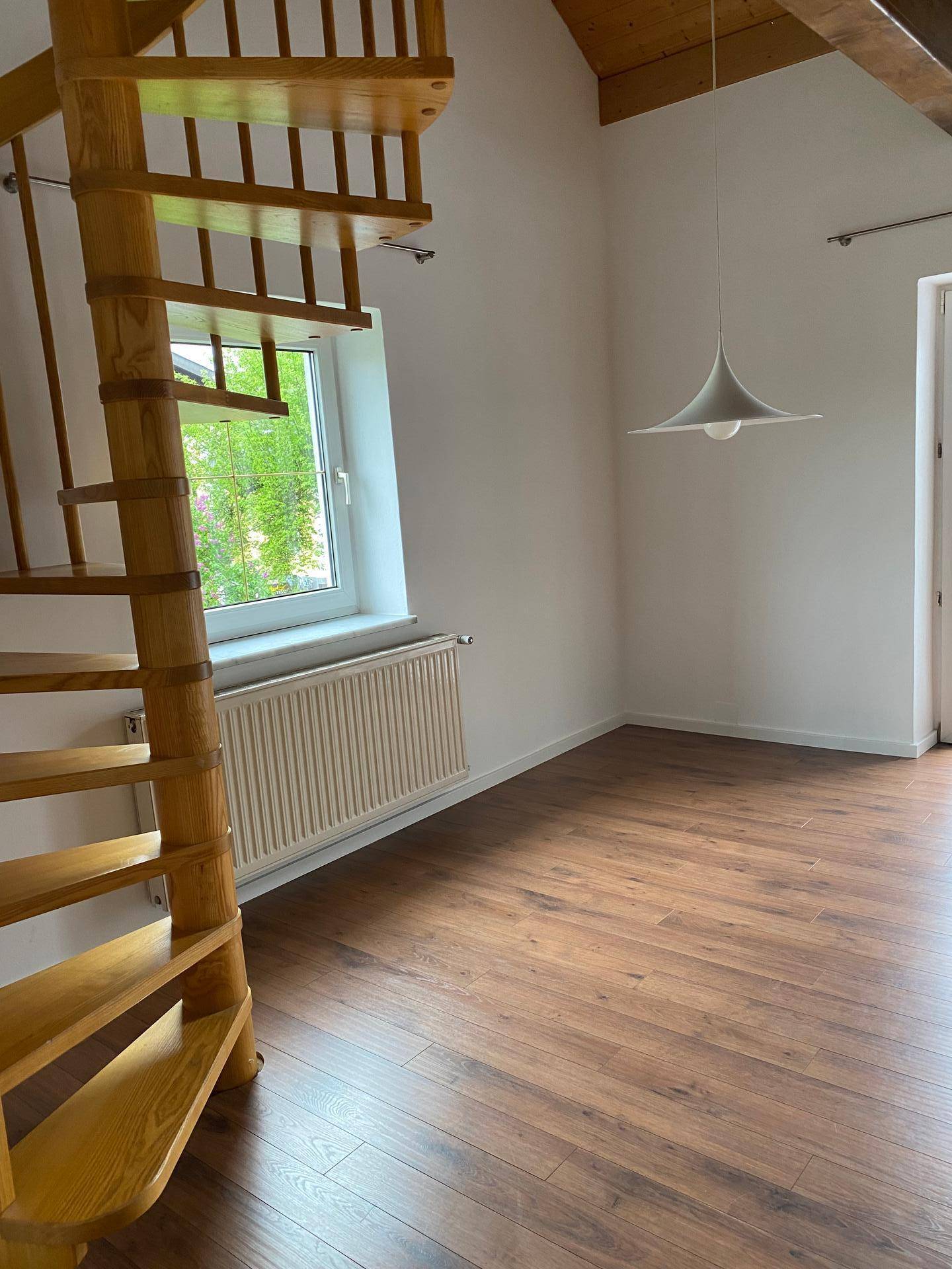 Lichtdurchfluteter Raum mit Wendeltreppe, Holzbalken und Blick ins Grüne durch das Fenster.