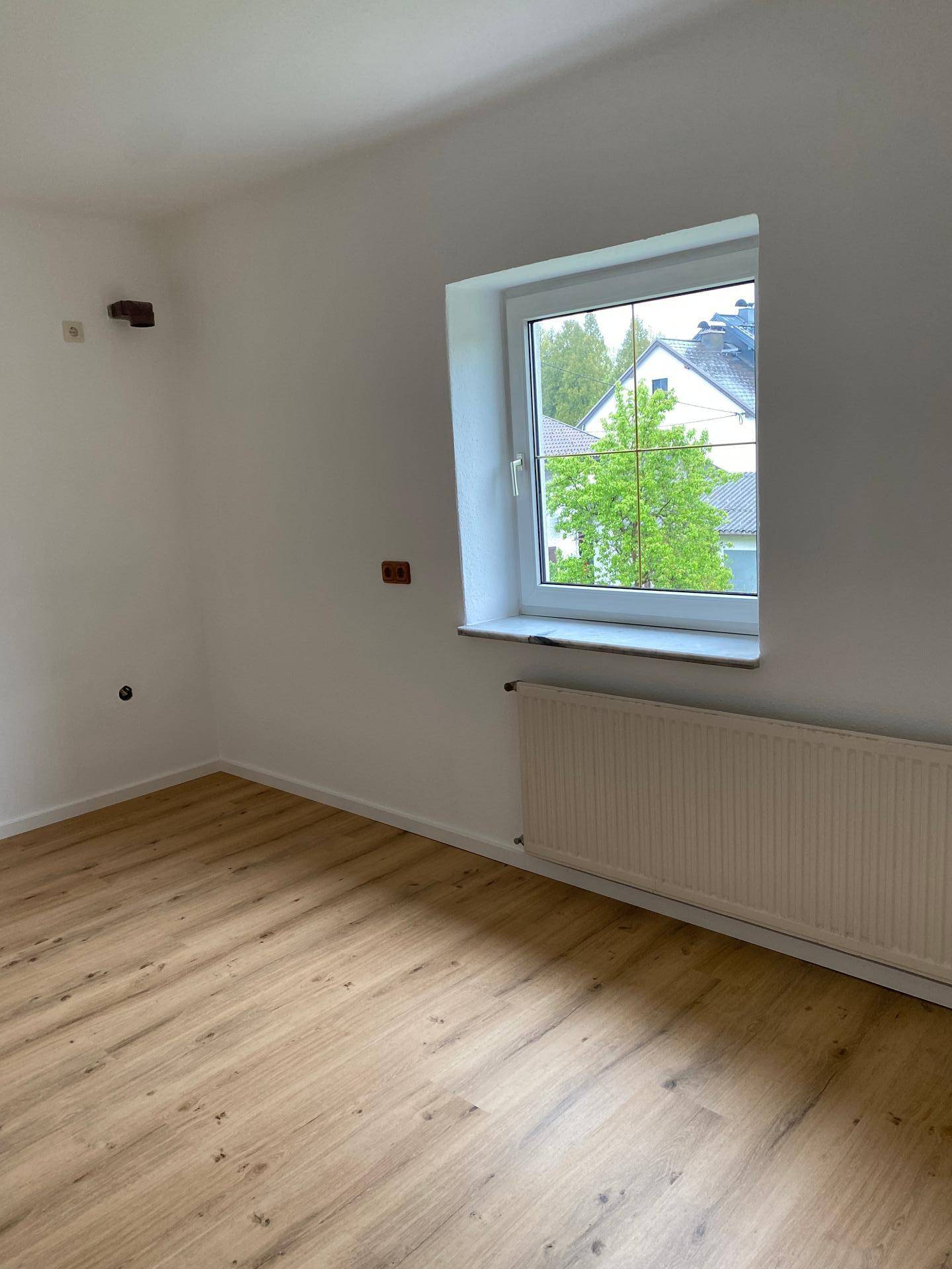 Helles Schlafzimmer mit Laminatboden, Heizkörper und Fenster mit Blick ins Grüne.