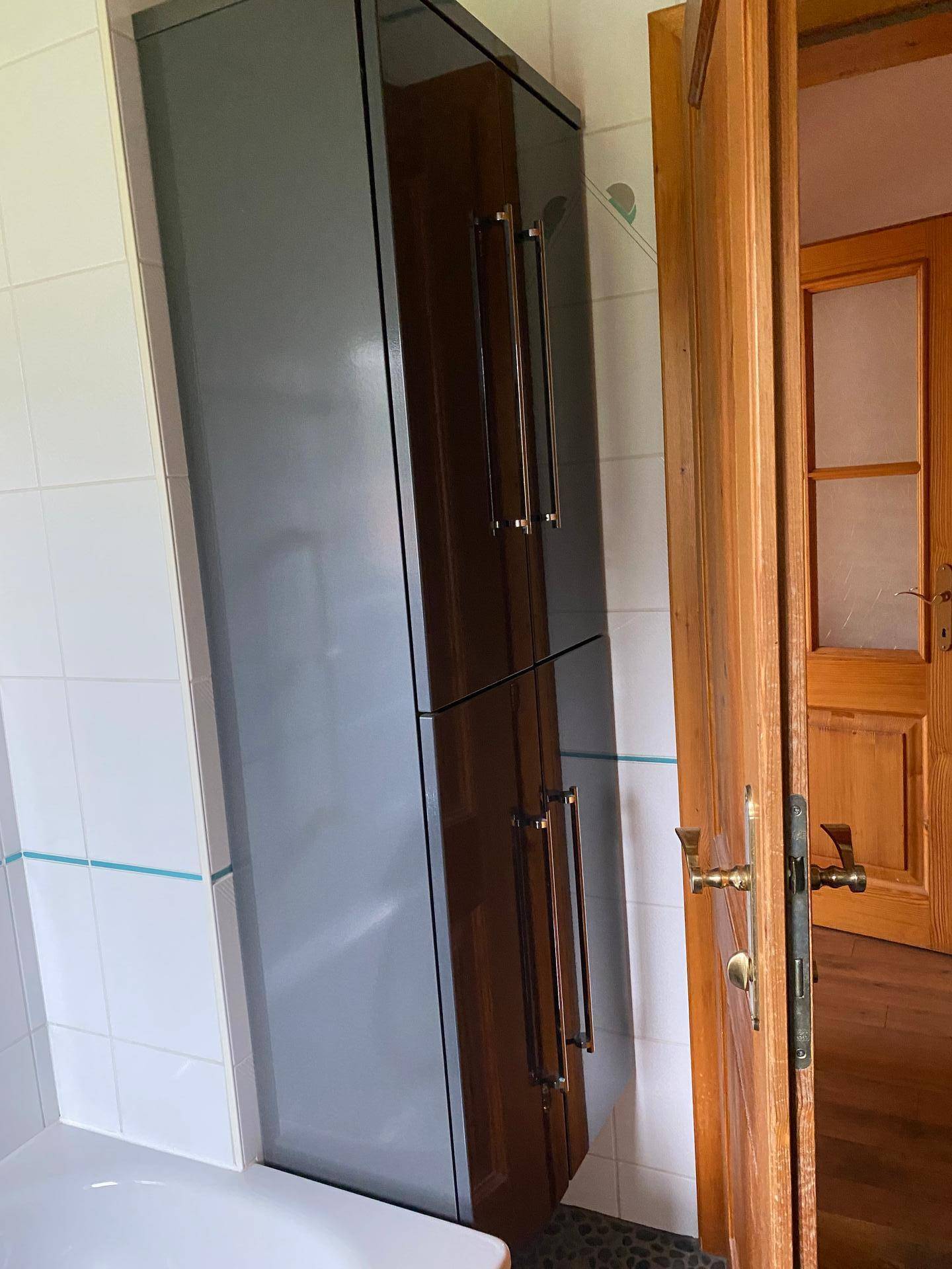 Badezimmerschrank mit dunklen Hochglanzfronten neben einer Holztür.