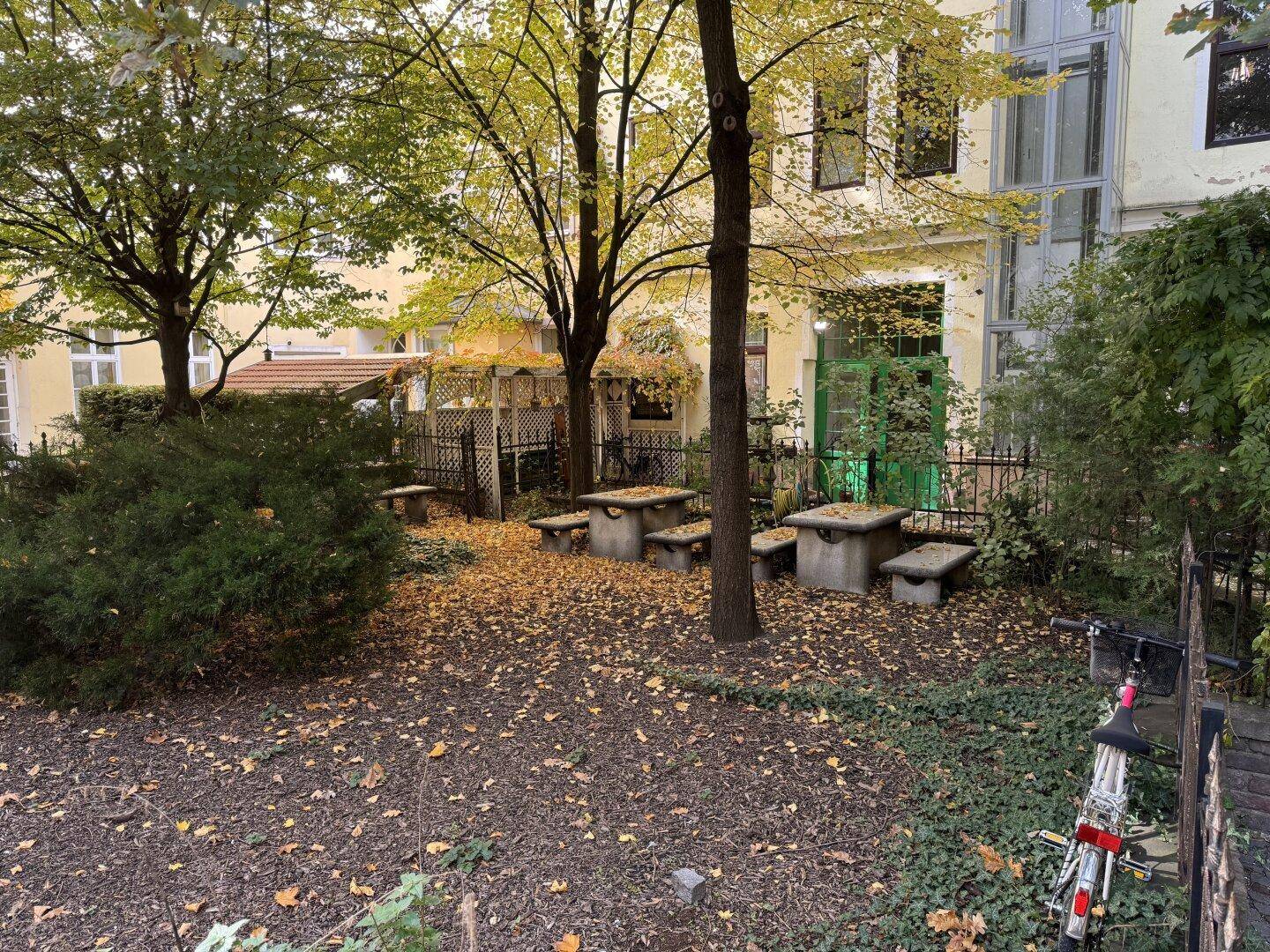 Begrünter Innenhof mit Bäumen, herbstlichem Laub und Steintischen für gemeinsame Nutzung.