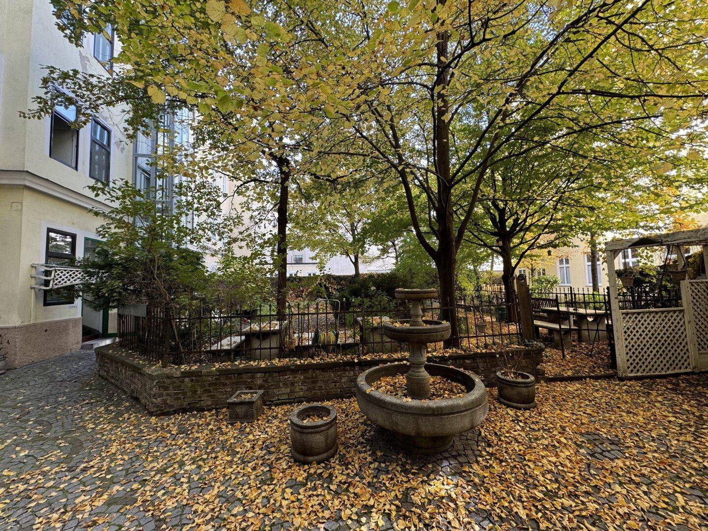 Idyllischer Innenhof mit Herbstlaub, hohen Bäumen und einem klassischen Steinbrunnen.