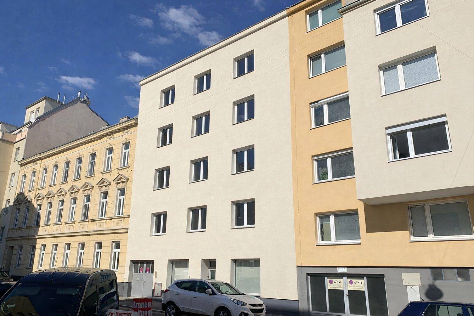 Ansicht der Gebäudefassade von der Straße mit parkenden Autos und angrenzenden Altbauten.