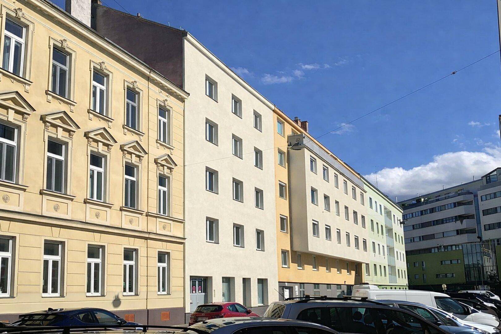 Weitläufige Ansicht der Gebäudefassade mit angrenzenden Gebäuden und parkenden Fahrzeugen unter blauem Himmel.