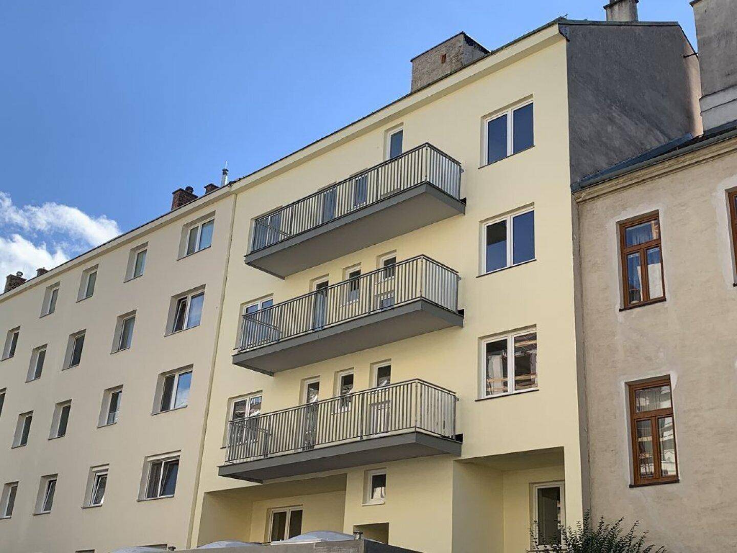 Schräge Ansicht der Gebäudefassade mit Balkonen und angrenzendem Altbau unter blauem Himmel.