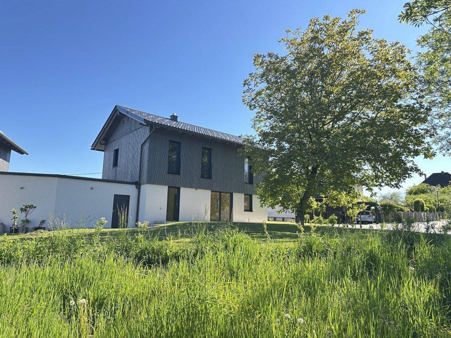 Neubau mit Holzverkleidung und weißer Fassade, umgeben von grüner Wiese und Bäumen.