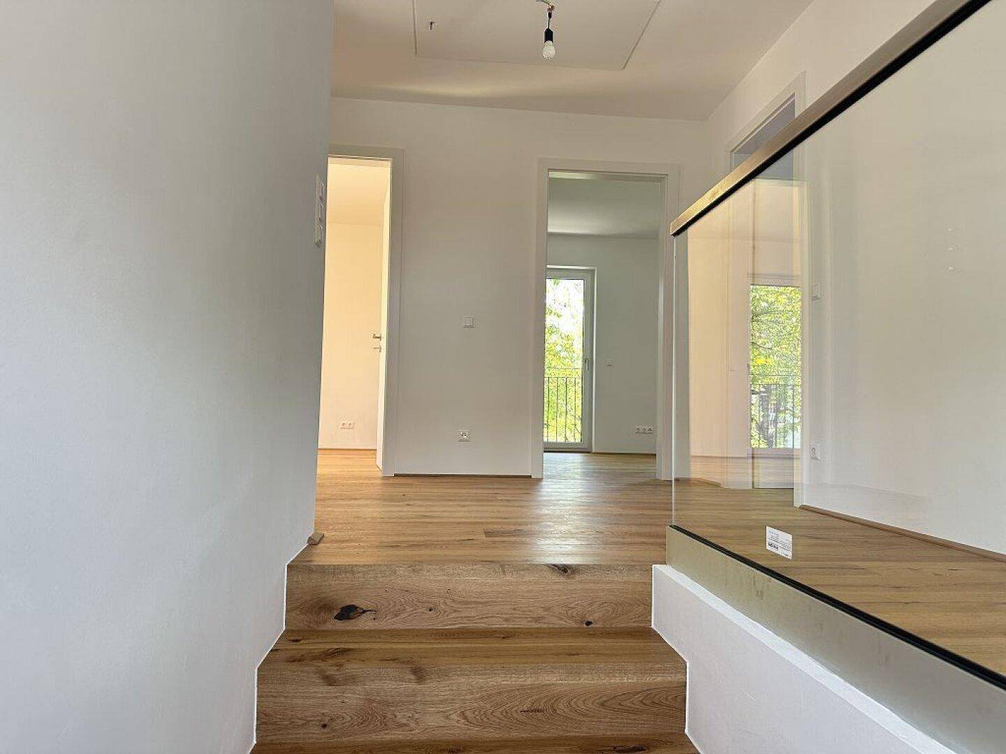 Heller Flur mit Holztreppe und Glasgeländer, der zu weiteren Räumen führt.