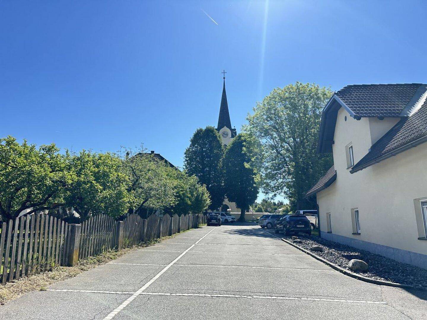 Ländliche Straße mit parkenden Autos und einer Kirche im Hintergrund unter blauem Himmel.