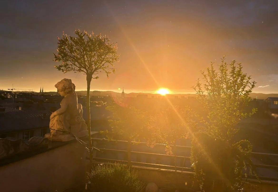 Atemberaubender Sonnenuntergang von einer Terrasse mit einer Statue und jungen Bäumen.