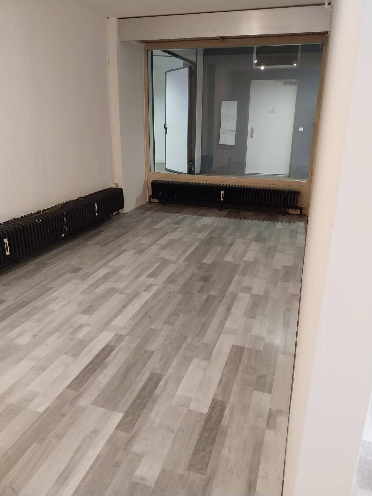 Gewerbefläche mit modernem Dielenboden, weißen Wänden und dunklen Heizkörpern unter einem großen Schaufenster.