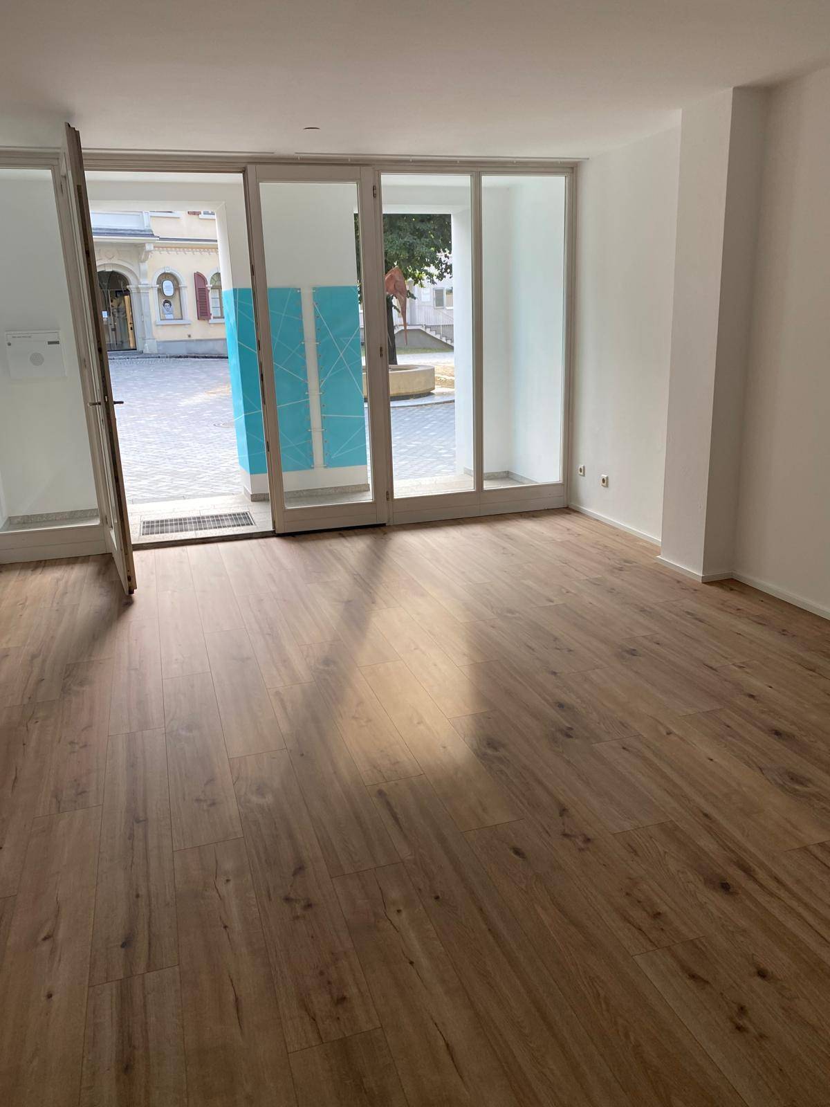 Heller, leerer Raum mit großen Glasfronten, Laminatboden und Blick auf einen gepflasterten Innenhof.