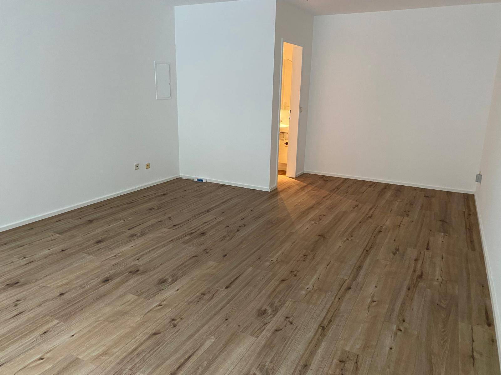 Leerer Raum mit weiß gestrichenen Wänden, hellem Laminatboden und Zugang zu einem angrenzenden Badezimmer.