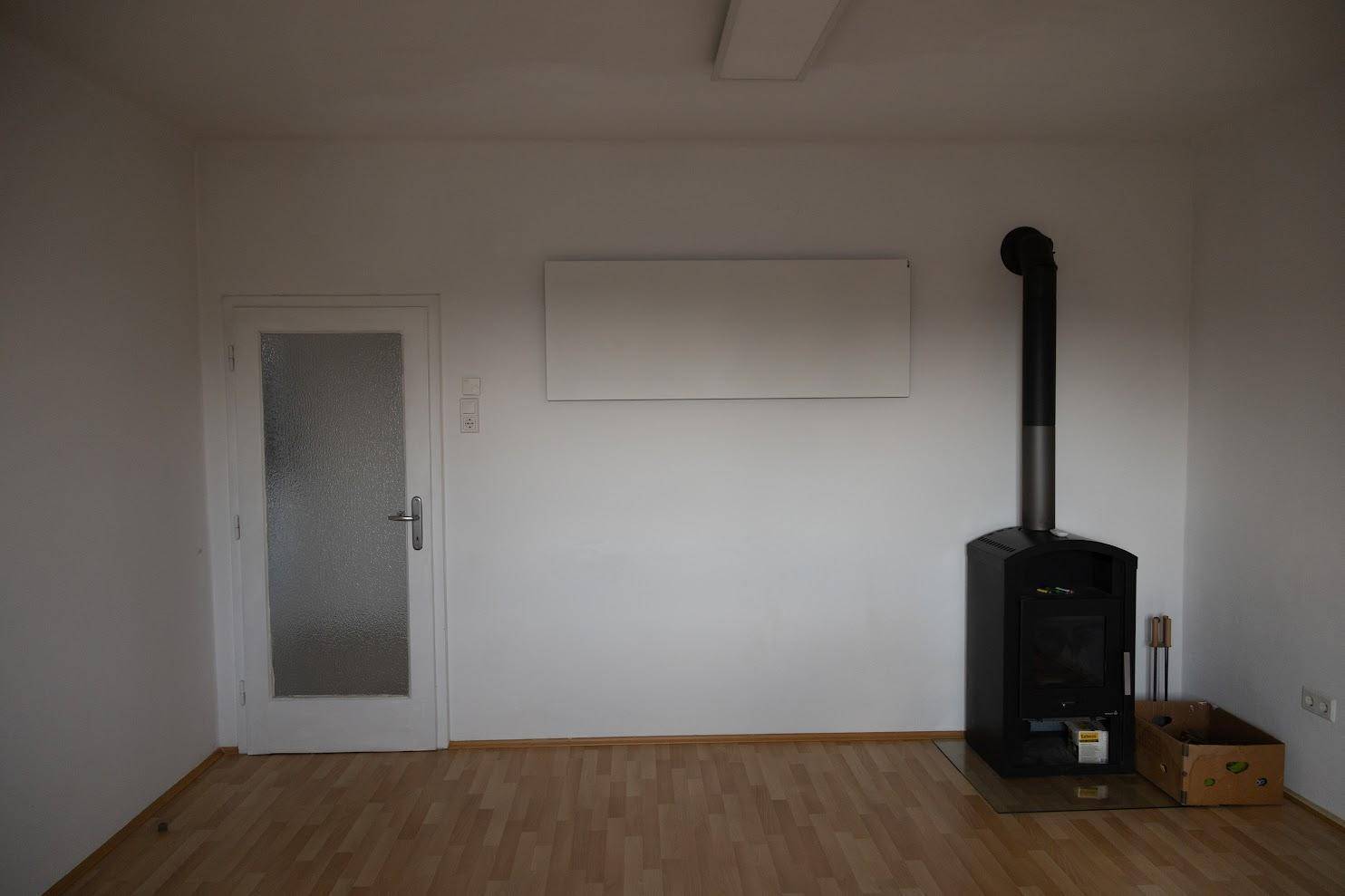 Helles Zimmer mit Holzboden, einem modernen Kaminofen und einer Tür mit Glaseinsatz.