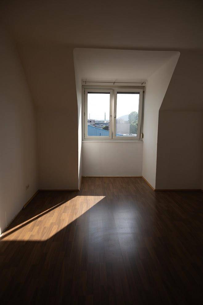 Sonniges Dachgeschosszimmer mit dunklem Holzboden und einem Fenster, das viel Licht hereinlässt.