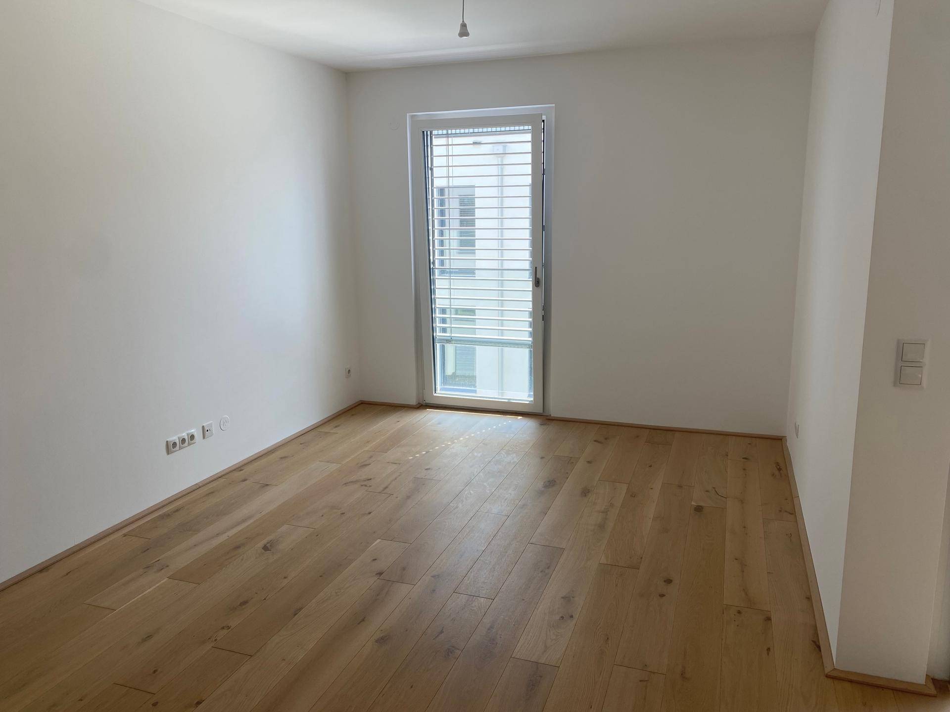 Leeres Zimmer mit hellem Parkettboden, weißen Wänden und großem Fenster mit Jalousien.