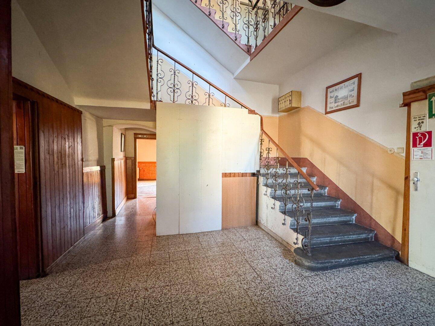 Geräumiger Eingangsbereich mit Terrazzoboden und geschwungener Treppe, zeigt Renovierungsbedarf.