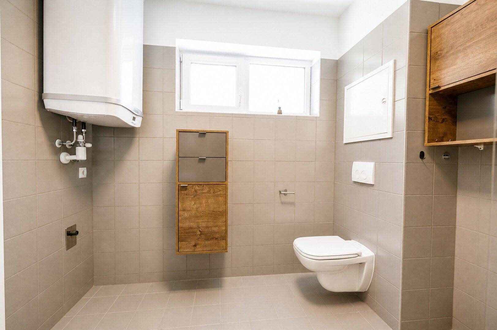 Separates WC mit wandhängender Toilette, Fenster und modernen Fliesen in Beige und Grau.