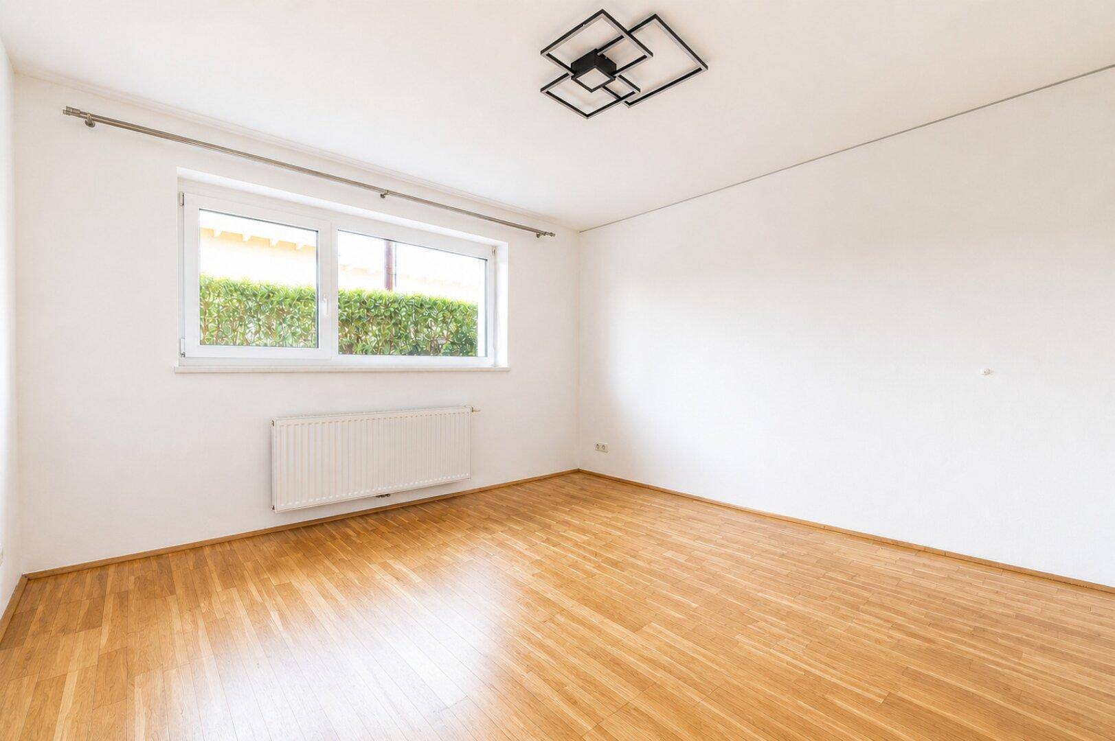Helles Zimmer mit Parkettboden, Heizkörper und Blick auf die grüne Hecke im Außenbereich.